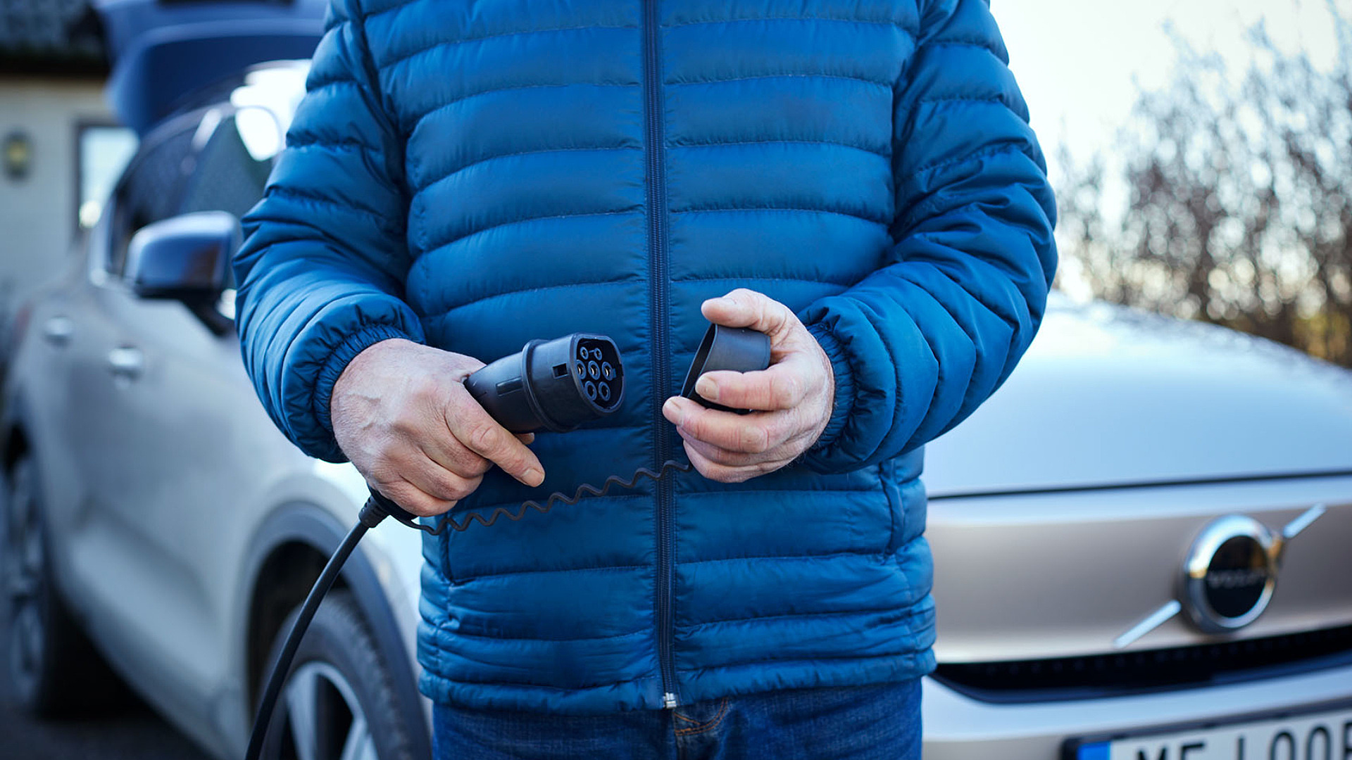 Man i blå jacka håller i en laddkabel för att sätta igång laddningen av sin elbil.